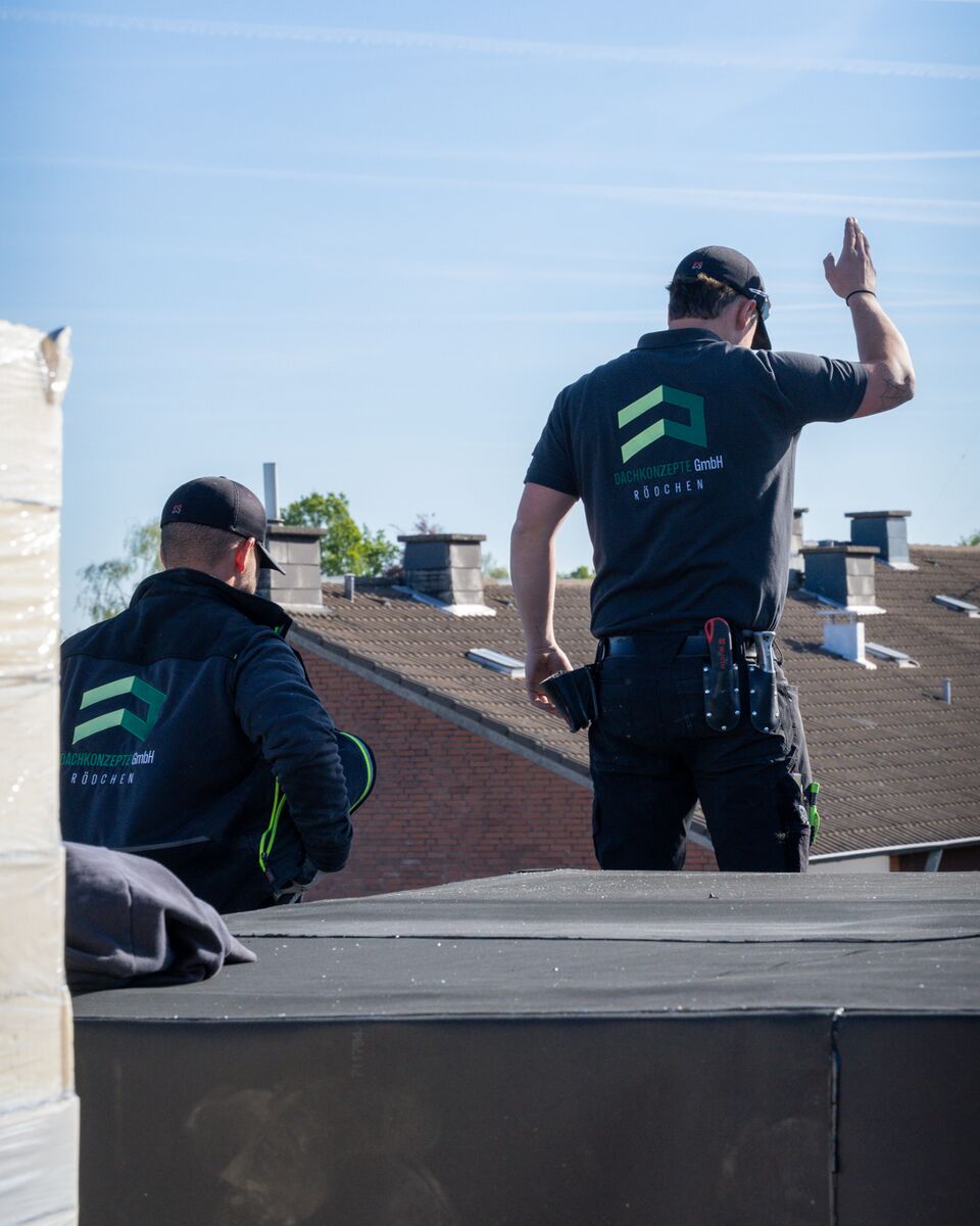 Zwei Dachdecker von DACHKONZEPTE auf einem Flachdach, einer gibt dem Kranführer ein Handzeichen
