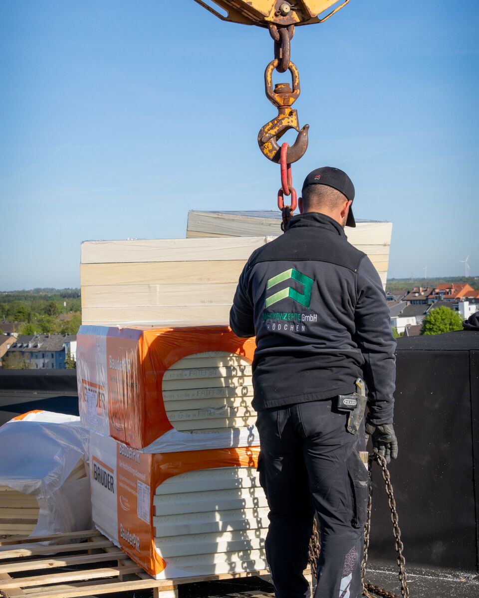 Dämmplatten hängen am Kranhaken über einem Flachdach, ein Dachdecker von DACHKONZEPTE nimmt sie an